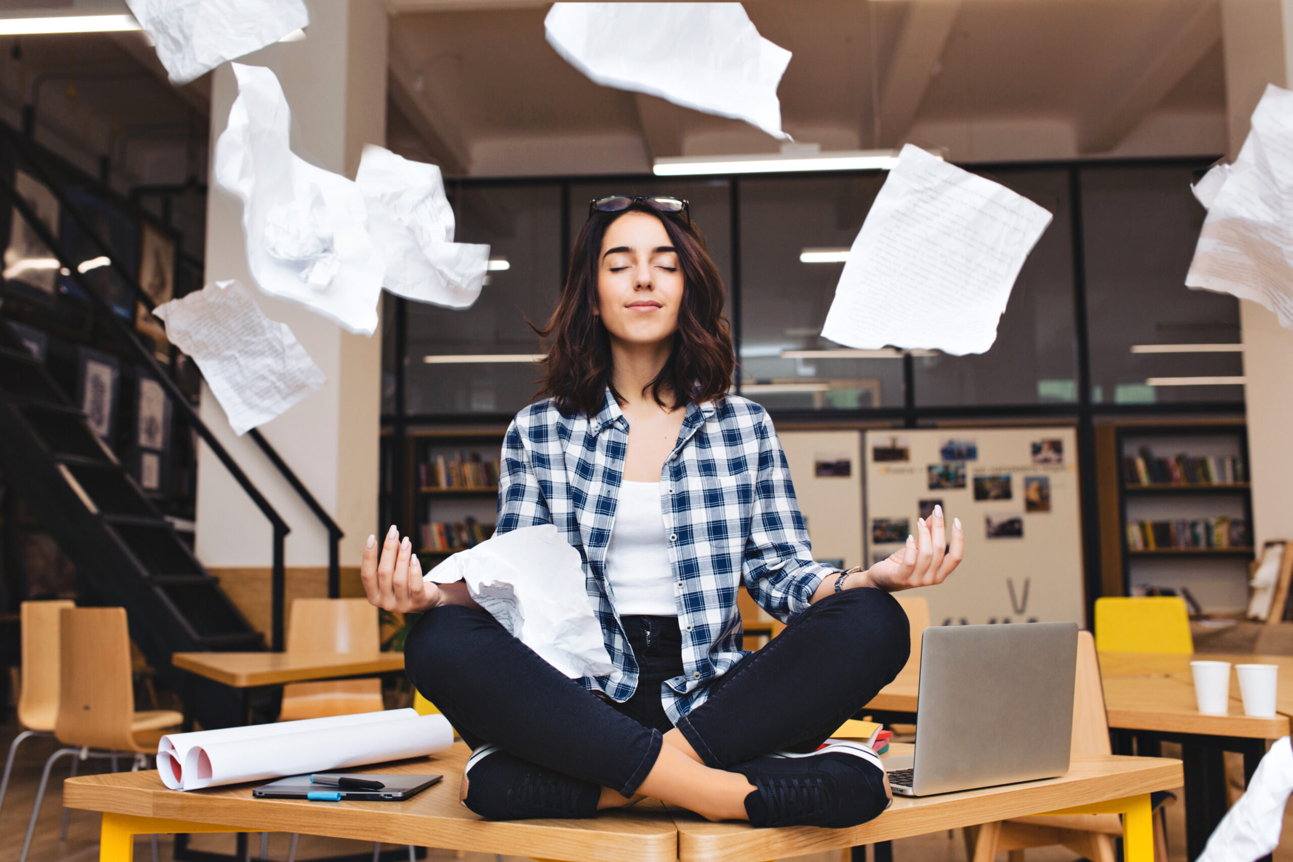 young pretty joyful brunette woman meditating on table surround work stuff and flying papers. cheerful mood, taking a break, working, studying, relaxation, true emotions.